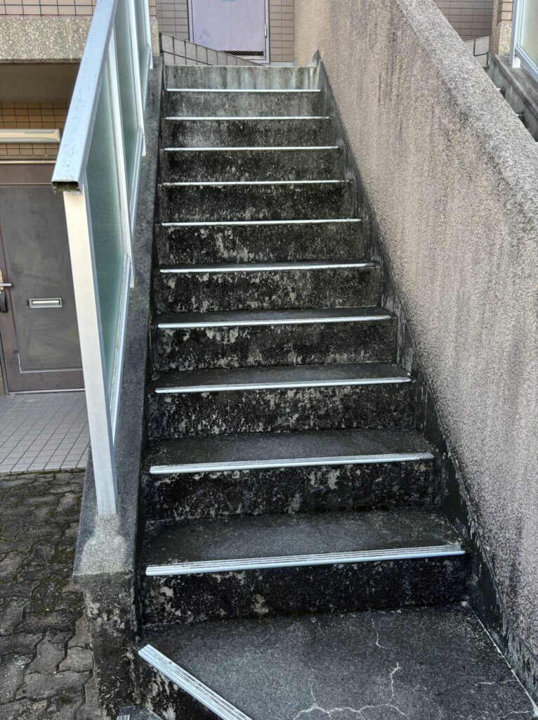 大阪府豊中市のマンション共用階段の清掃前。長年の雨だれや黒カビ、苔が固着して全体が真っ黒に変色し、古びた印象を与えるコンクリート階段の状態