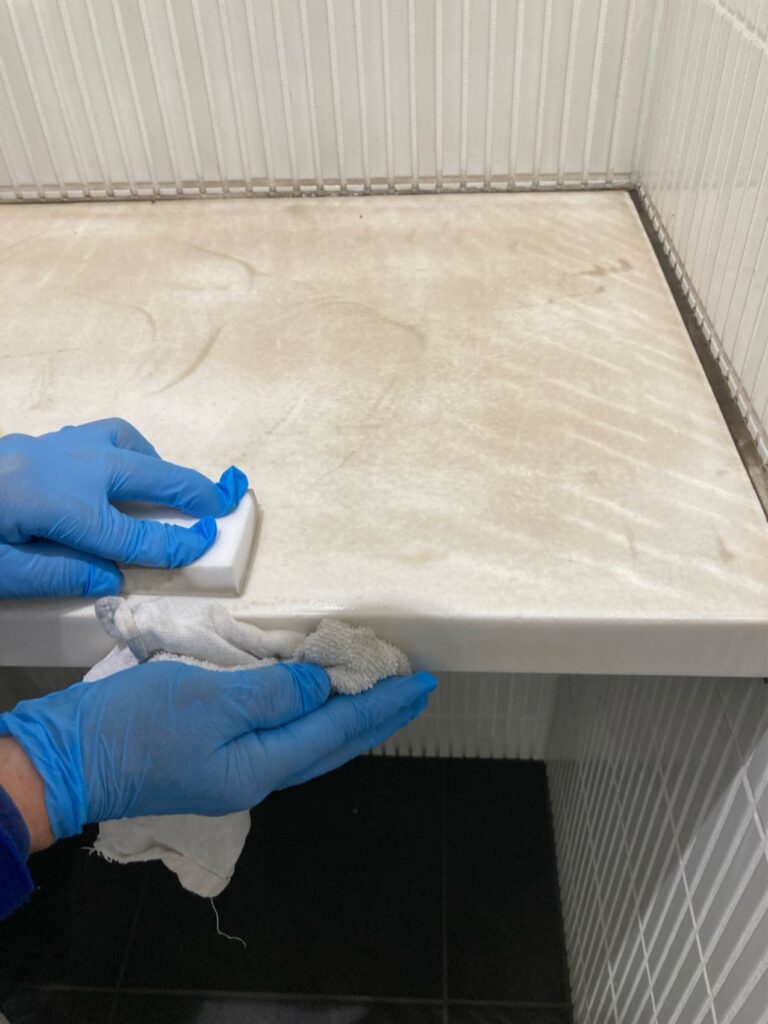 京都市の地下施設にて、トイレ個室の黒ずんだ荷物置きを特殊洗浄している作業風景。張り替えずにプロの技術で手垢汚れを丁寧に落としている様子