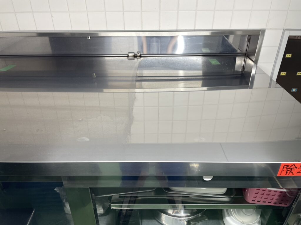 京都市の施設における調理室ステンレス作業台の清掃ビフォーアフター画像。左半分は毎日の使用による水垢や油汚れでくすんでいるのに対し、プロの拭き上げ清掃を行った右半分は汚れが完全に落ち、壁の白いタイルが鏡のように反射して映り込んでいる様子