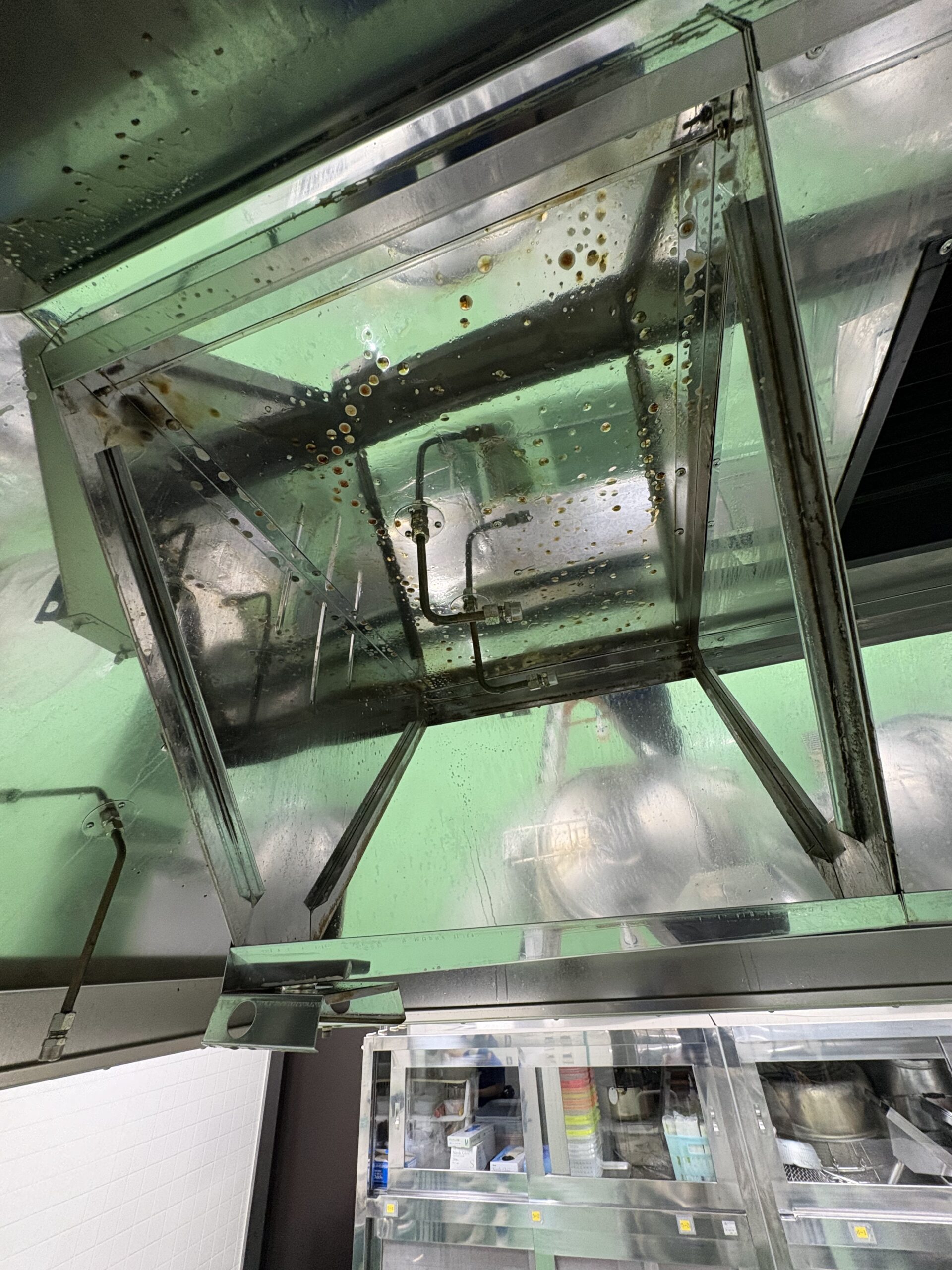 施設の厨房にある高所換気扇フードの内部。日々の清掃が届かないため、気化した黄色い油汚れが水滴のように無数にこびりつき、今にも落下しそうな不衛生な状態