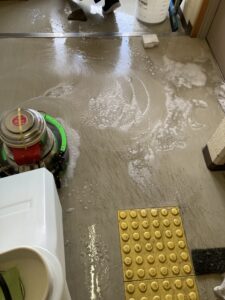 高齢者施設の共有スペースの床を、専用のポリッシャー（床洗浄機）と洗剤を用いて本格的に洗浄している作業風景。日常清掃では落ちない黒ずみや古いワックスを泡と一緒に浮き上がらせている様子