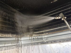 業務用の天井カセット型エアコンの熱交換器（アルミフィン）を、専用の高圧洗浄機でクリーニングしている様子。長年蓄積したカビや繊維ボコリが真っ黒な汚水となって大量に流れ落ちている作業風景